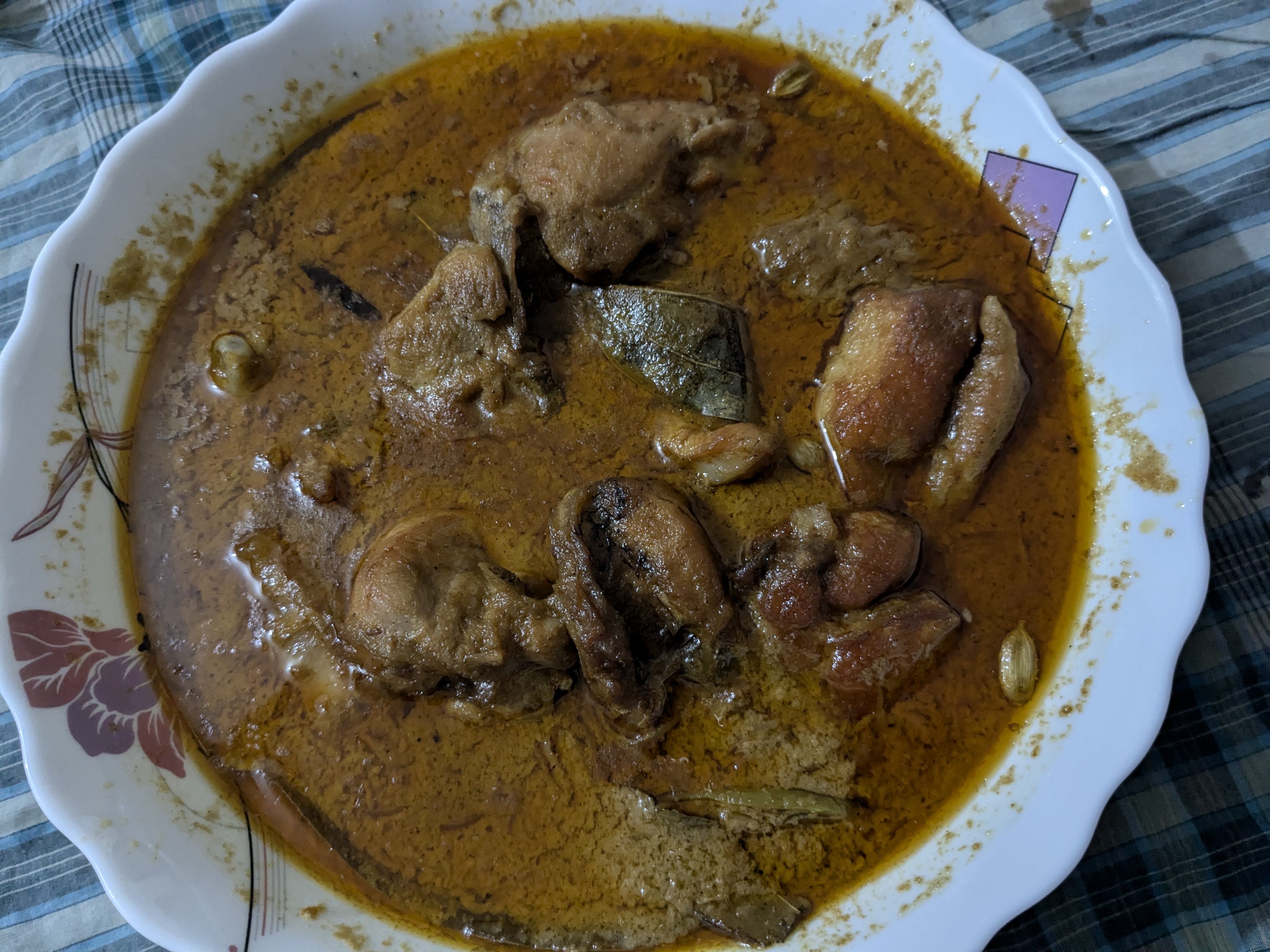 A white plate with red floral patterns holds a rich, spicy chicken curry. The dish features hearty chunks of chicken in a thick, reddish-brown sauce.