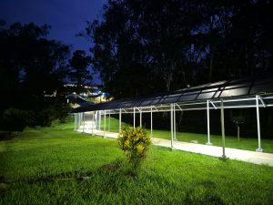 A brightly lit covered walkway with a curved translucent roof stretches through a dark, grassy park at night, leading toward buildings visible in the distance.
