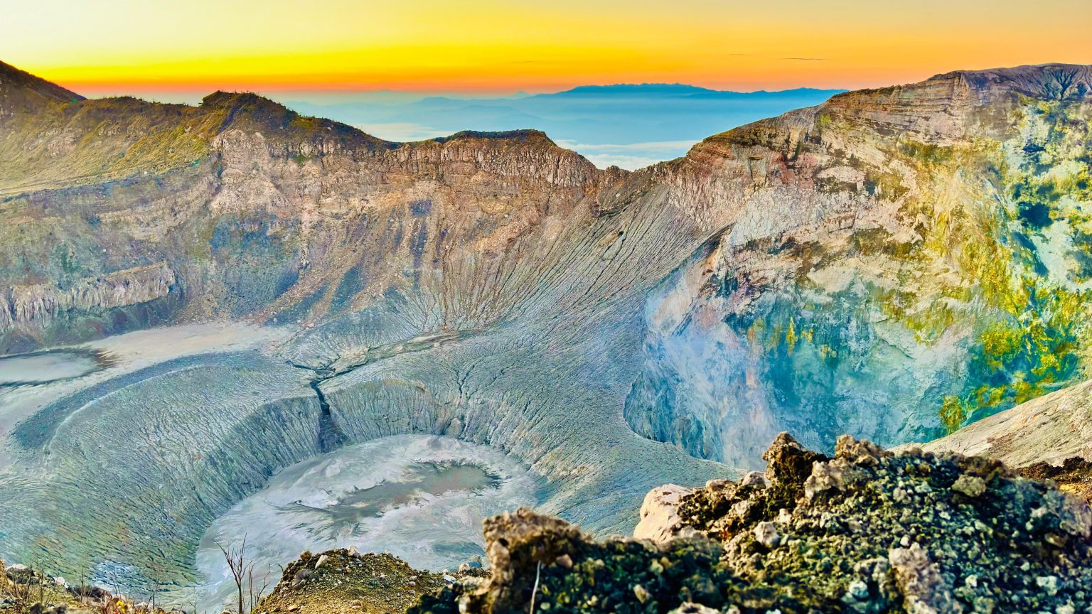 A broad volcanic crater with rugged mineral-coloured walls beneath a golden horizon.