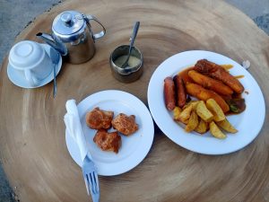 A concrete table set, painted to appear wooden set with breakfast, including pastries, sausages, potato wedges, and a tea set.
