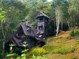Casa de madera con una torre, rodeada de árboles, vegetación y una ladera con flores.
