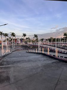 View of a multi-level go-kart track with winding lanes, elevated ramps, and safety barriers. Palm trees, colorful flags, and modern light poles surround the circuit beneath a wide blue sky with scattered clouds during the late afternoon.