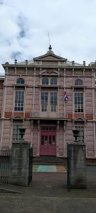Front facade of the Escuela Metálica (Metal School), built in 1890 in San José, Costa Rica, featuring pink prefabricated iron panels, arched windows, an ornate wrought-iron balcony and the national coat of arms above the entrance.