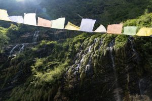 Colorful prayer flags strung across a rocky moss-covered cliff with multiple small waterfalls