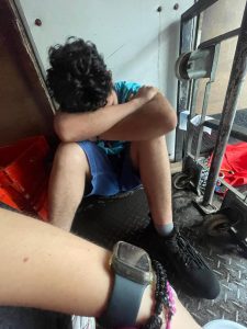 High-angle shot of a person with dark curly hair sitting on a black diamond-plate floor, resting their head on their crossed arms. They are wearing a blue t-shirt, blue shorts, and black sneakers. In the foreground, a wrist with a smartwatch is partially visible. A metal cart and a red container are in the background against a white wall.
