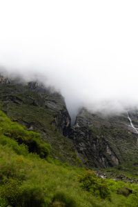Rocky mountain gorge with green vegetation and a distant waterfall, upper slopes covered in thick fog