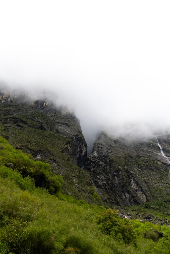 Rocky mountain gorge with green vegetation and a distant waterfall, upper slopes covered in thick fog