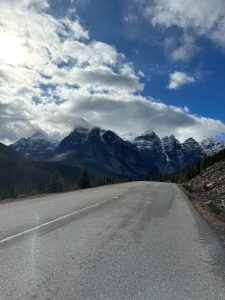 A mountain road leading toward snow-covered peaks under a blue sky with large white clouds.