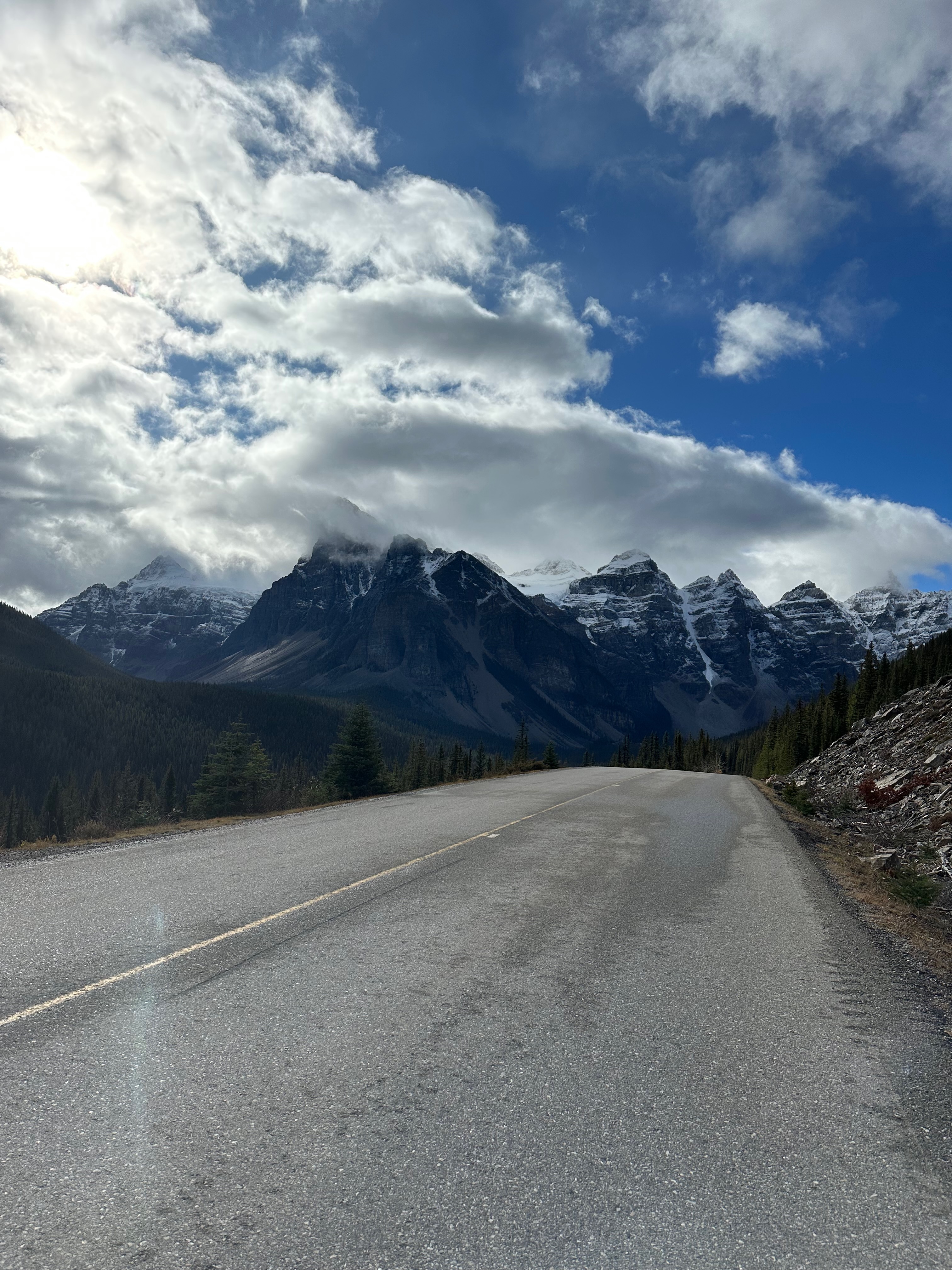 A mountain road leading toward snow-covered peaks under a blue sky with large white clouds.