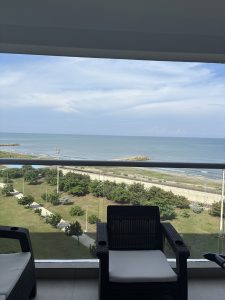 The image shows a serene view from a balcony overlooking a calm sea. In the foreground, there are two black wicker chairs with light-colored cushions positioned on a tiled floor. The balcony railing features a glass panel, offering an unobstructed view.
