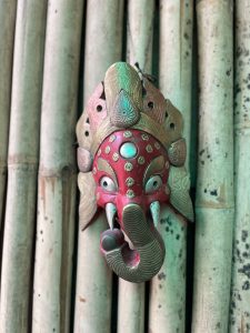 A red and brass decorative mask of the Hindu deity Ganesha hangs mounted against vertical bamboo stalks. The elephant-headed mask features a vibrant red face decorated with metallic brass emblems, large round silver eyes, small white tusks, a curved brass trunk, and an ornate, fan-like metallic crown.