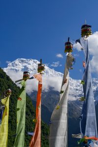 Tall vertical prayer flags in yellow, green, orange, white, and blue on wooden poles with a snow-capped mountain and forested hillside in the background