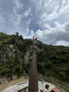 A high-angle view shows a tall, slender dark monument tapering upward with a red flag flying near its top. It stands on a concrete platform beside a muddy river. Lush green, rocky mountains rise sharply in the background under a cloudy blue sky with large white clouds.
