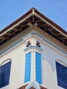 The corner of a traditional Kerala house features blue-painted pillars, wooden roof beams, and decorative carvings beneath the tiled roof. Captured at Mangode, Puthunagaram, Palakkad.