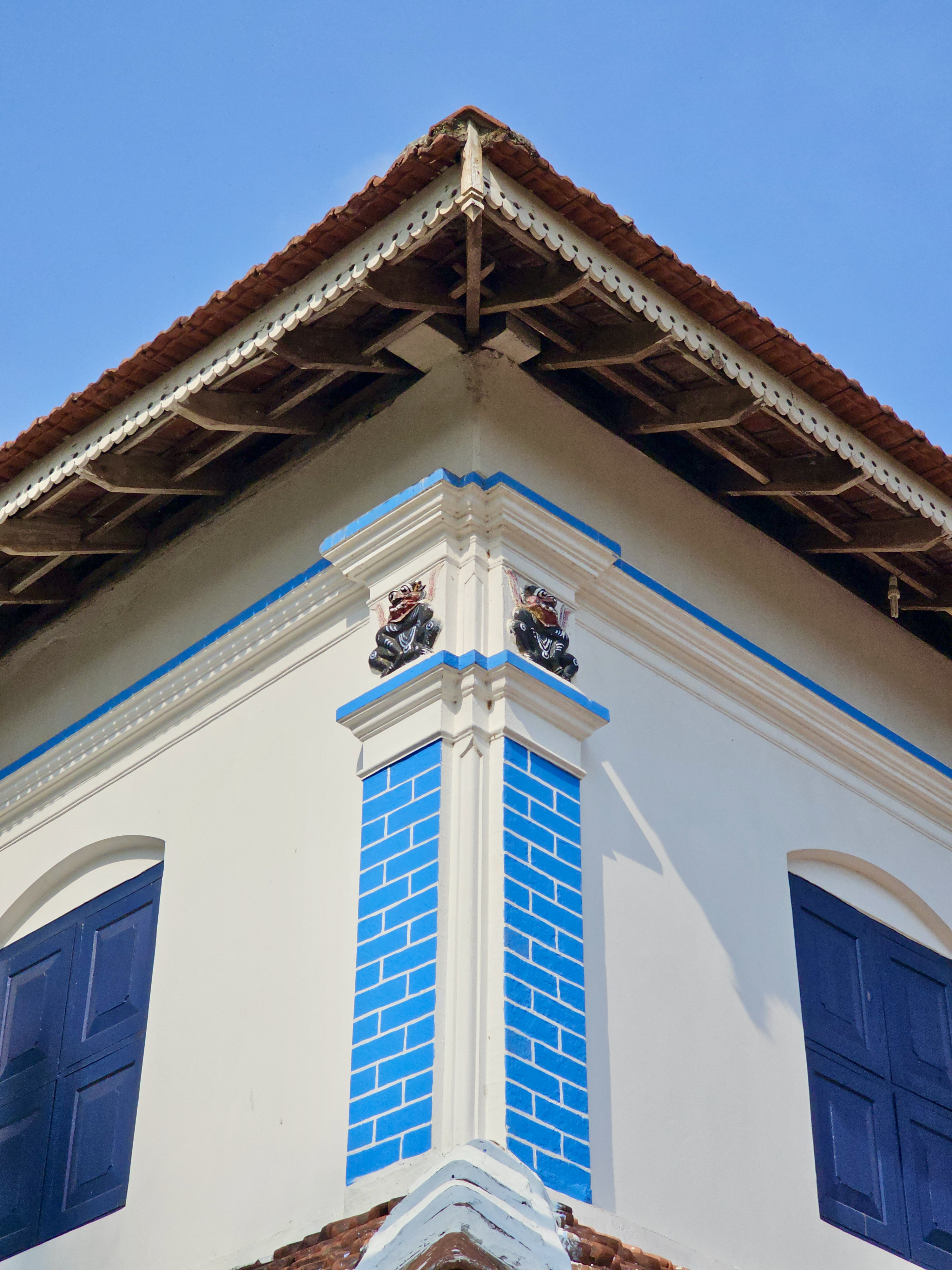 The corner of a traditional Kerala house features blue-painted pillars, wooden roof beams, and decorative carvings beneath the tiled roof. Captured at Mangode, Puthunagaram, Palakkad.