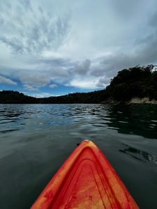The bow of a kayak with a hull fading from reddish-orange to yellow stretches toward calm water that reflects a cloudy sky dotted with wispy clouds and patches of blue