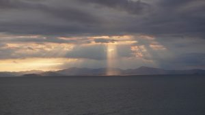 Sunlight breaks through dark clouds over a calm body of water, illuminating distant mountains on the horizon.