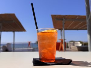 Orange drink with ice, orange slice, and straw on a white table. Blue sky, shaded structures, and ocean in background.