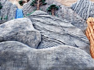 Artificial rock formation diorama with textured gray boulders, a painted blue waterfall, miniature bonsai-style trees, and an orange stalactite-like structure