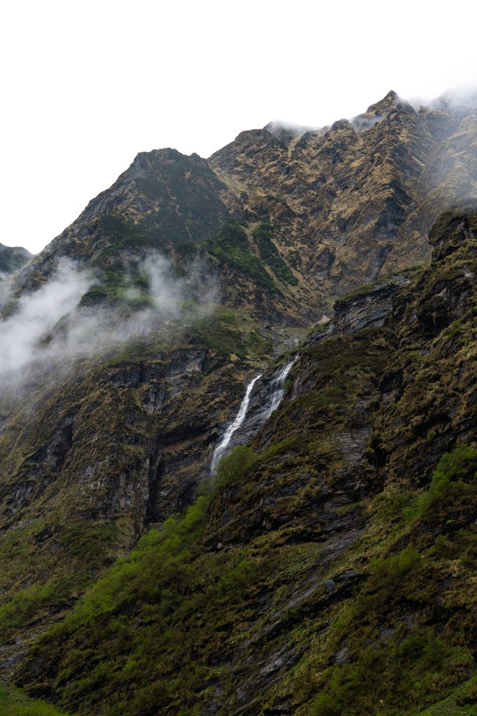 Thin waterfall streaming down a steep mountainside with rocky peaks and drifting clouds