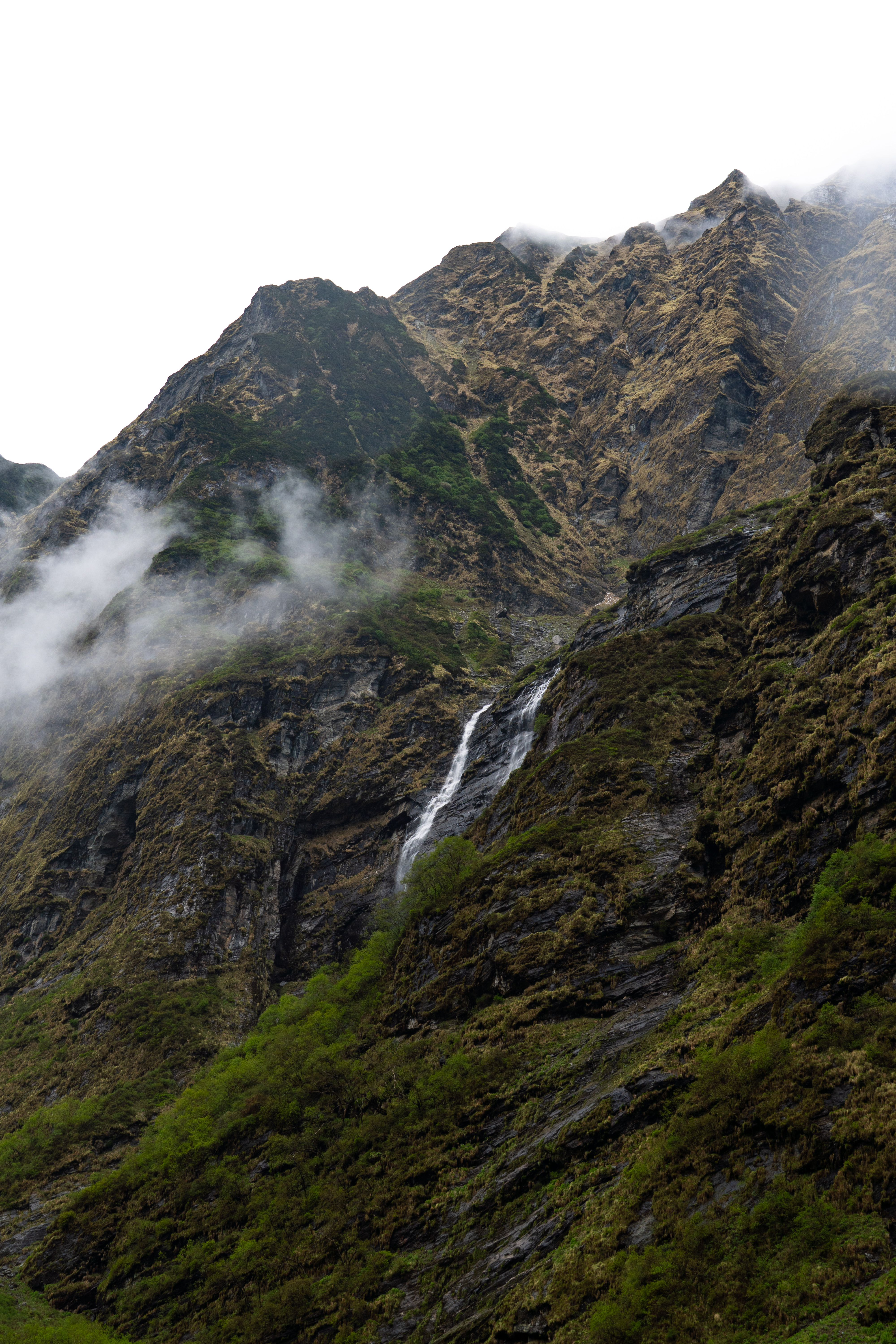 Thin waterfall streaming down a steep mountainside with rocky peaks and drifting clouds