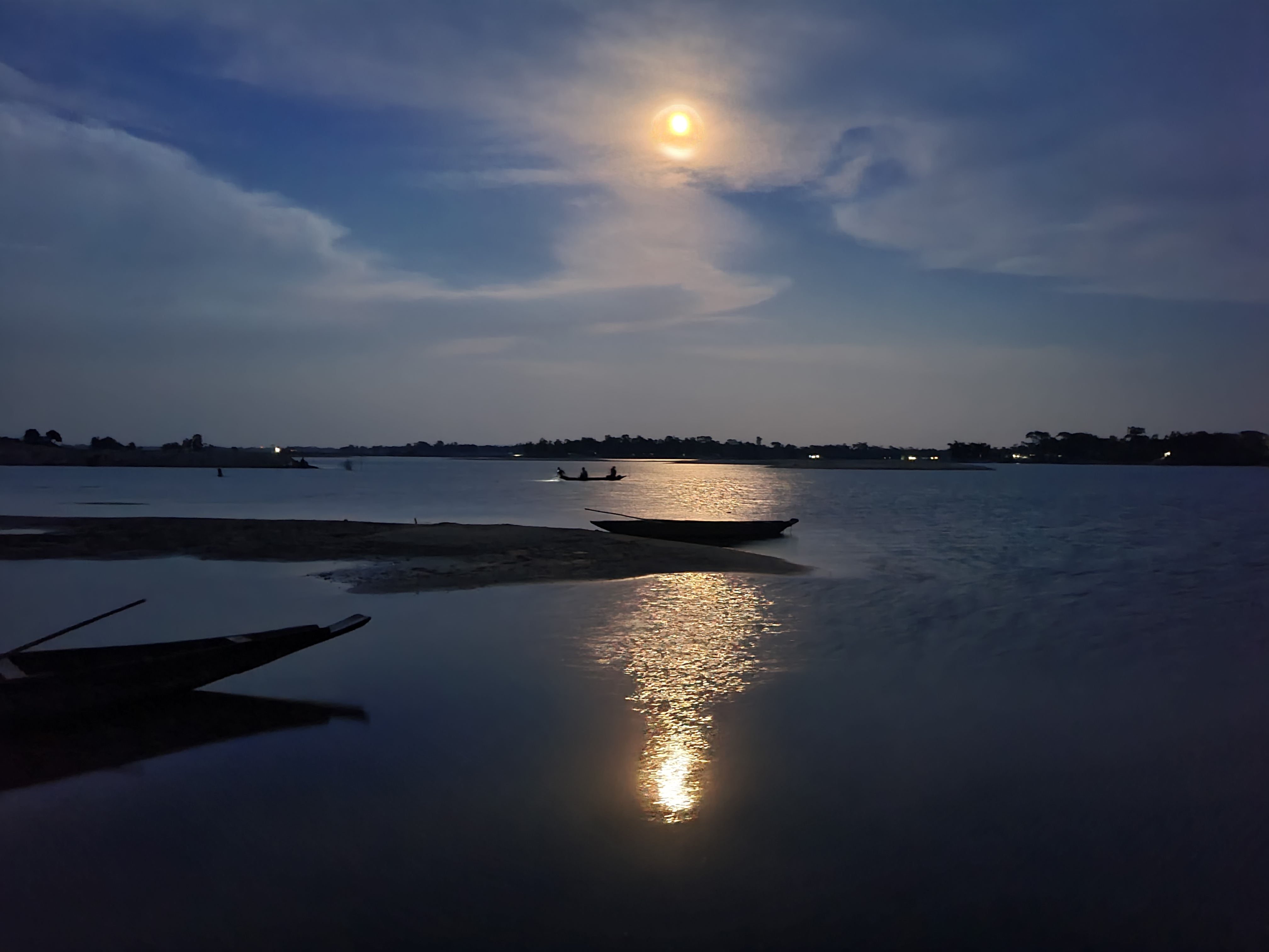 Moonlight reflects off the calm water of a river where two wooden boats are moored near the shore at dusk.