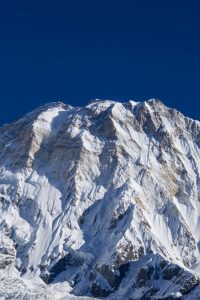 Massive snow and ice-covered mountain face with jagged rocky ridges against a deep blue sky