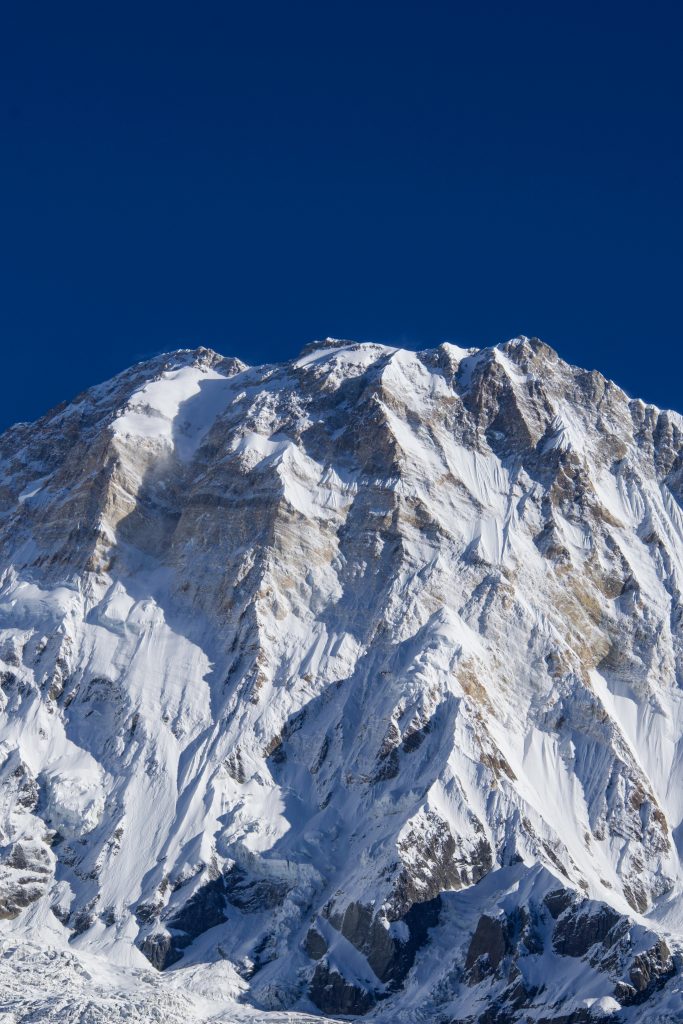 Massive snow and ice-covered mountain face with jagged rocky ridges against a deep blue sky