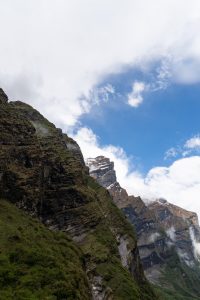Steep mountain cliffs with patches of green vegetation rise under a bright blue sky with scattered clouds
