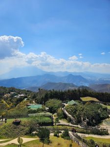 A panoramic view of mountain ranges under a bright blue sky with large white clouds, with gardens, buildings, and walking paths in the foreground