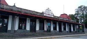 Side facade of the historic Ferro-Carril al Atlántico train station in Costa Rica, with a red roof, ornate wooden trim and a clock reading “1908” above the entrance.
