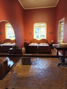 Historic colonial bedroom at the National Museum of Costa Rica in San José. Features wooden furniture with carved headboards, deep red walls, arched windows and original stone tile flooring.