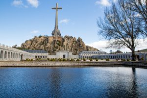 Panoramic view of Cuelgamuros Valley with a pond, large stone cross, basilica, and blue sky.