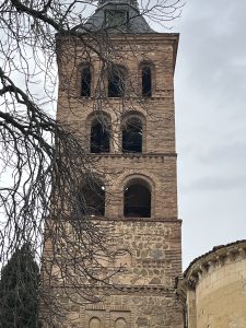 Mudéjar-style brick bell tower with arched openings and stone lower walls beneath a cloudy sky.
