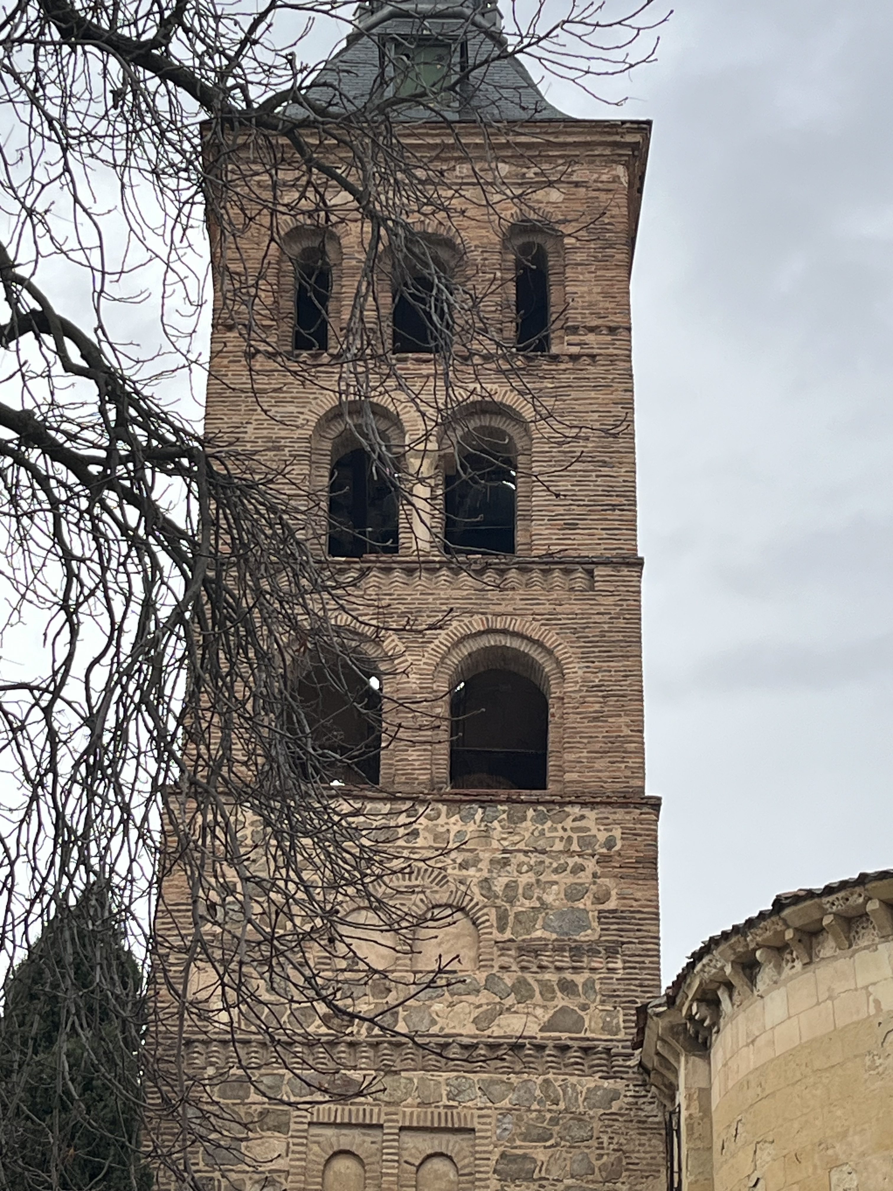 Mudéjar-style brick bell tower with arched openings and stone lower walls beneath a cloudy sky.
