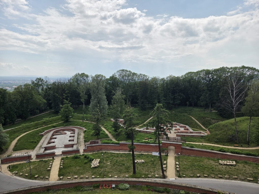 Aerial view of a park with the excavated foundations of old brick fortifications, surrounded by green lawns, gravel paths, and dense trees, with a cloudy sky above.