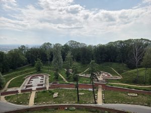 Aerial view of a park with the excavated foundations of old brick fortifications, surrounded by green lawns, gravel paths, and dense trees, with a cloudy sky above.

