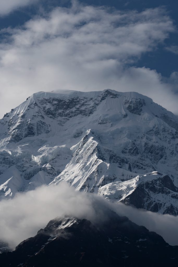 Massive snow-covered mountain with jagged rocky ridges, clouds gathered at its base and around the summit