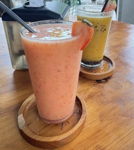 
Pink strawberry smoothie with a fresh strawberry slice beside a yellow seeded drink on a wooden table.