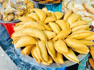 A plate filled with neatly arranged golden-brown fried pastries, likely empanadas or turnovers, displayed on a table among other wrapped snacks.