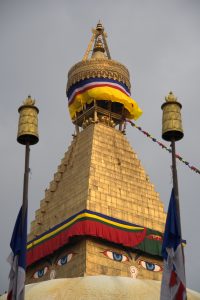 Golden stupa with colorful flags and Buddha's eyes painted on it under a gray sky, bordered by two prayer flags.
