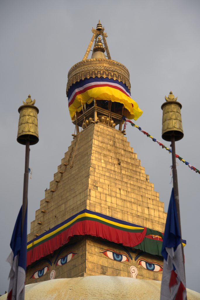 Golden stupa with colorful flags and Buddha’s eyes painted on it under a gray sky, bordered by two prayer flags.