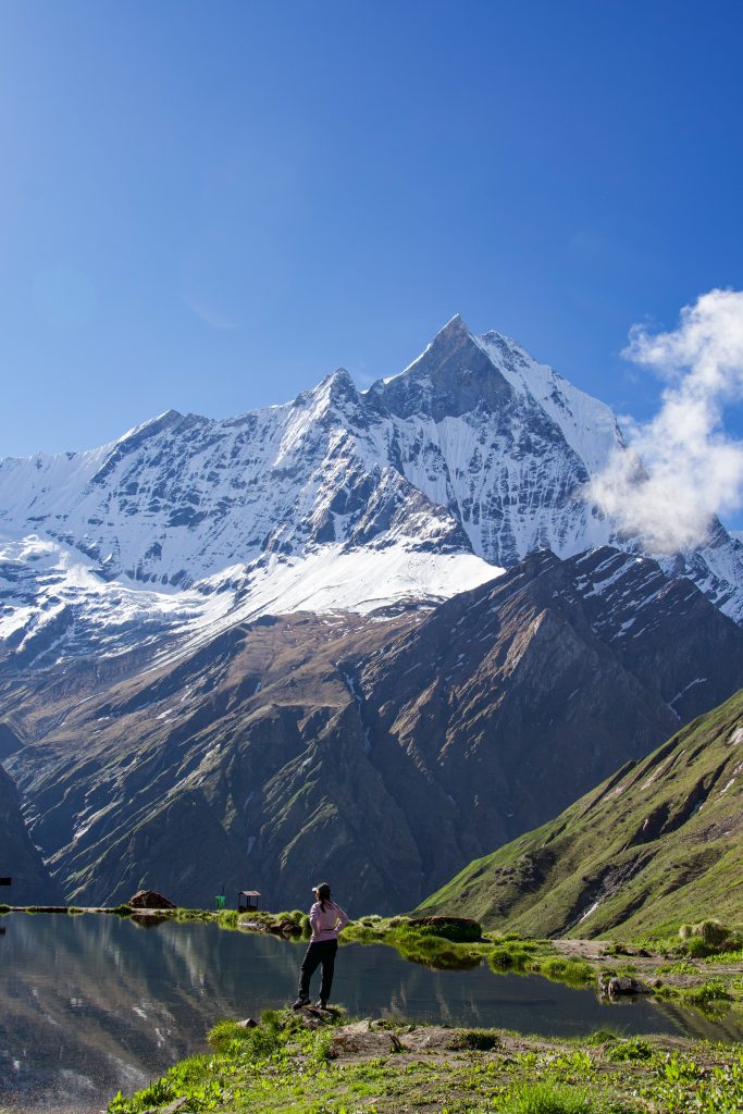 Person standing beside a still mountain lake facing a jagged snow-capped peak reflected in the water