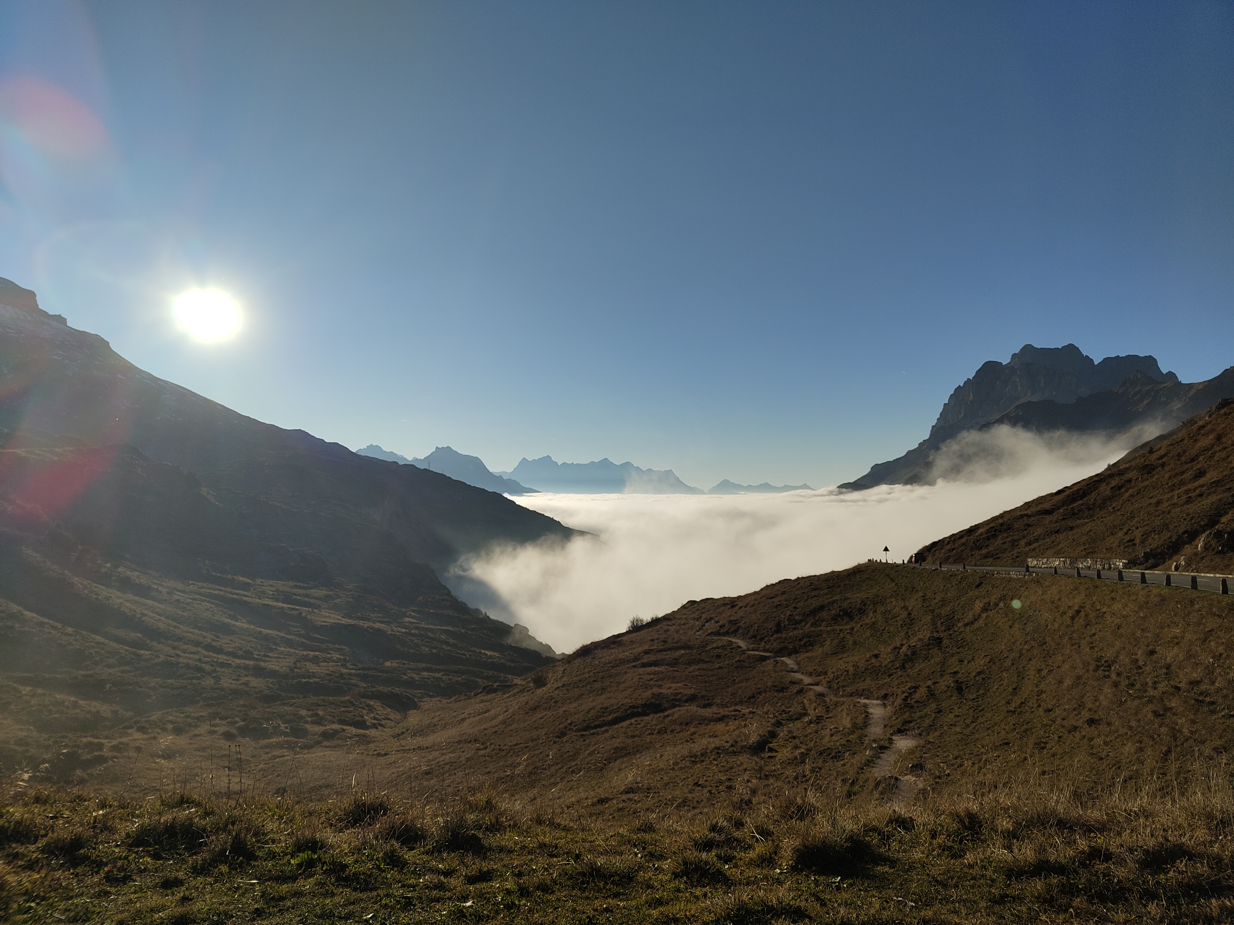 A high-altitude mountain valley looking down at a thick sea of white clouds nestled between peaks. The bright sun casts a lens flare over dry, grassy slopes in the foreground, with a narrow mountain road hugging the right ridge.