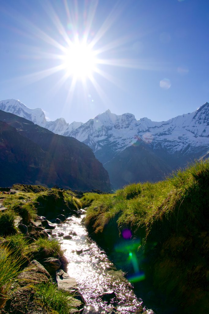 Sunlit stream running through a grassy valley with snow-capped mountains and bright sun in the sky