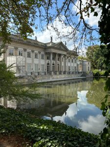 Historic building with columns beside a reflective pond, surrounded by greenery and partial scaffolding.