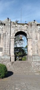 Stone entrance arch of the Santiago Apostle Ruins in Cartago, Costa Rica. A bronze bell hangs at the center above an ornate ironwork gate, with trees and open sky visible through the threshold.