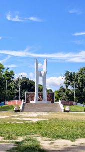 Rajshahi University Central Shaheed Minar standing under a bright blue sky, surrounded by green lawns, trees, and a wide open plaza.
