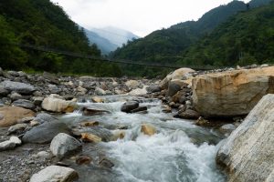Fast flowing river over boulders with a suspension bridge crossing between forested hills