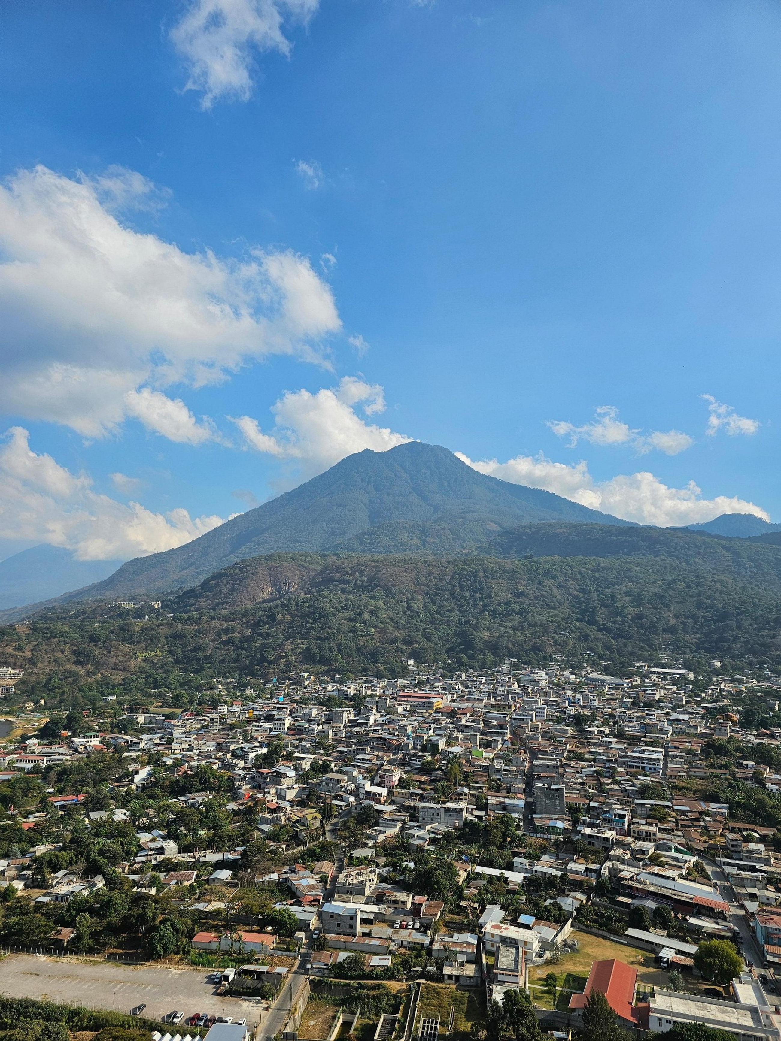 A town spread out at the base of a towering green volcano under a vivid blue sky with scattered clouds